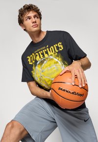 Sort grafisk t-shirt med teksten "Golden State Warriors" og et gult basketbolddesign. Grå shorts og en orange basketball holdt i hænderne.