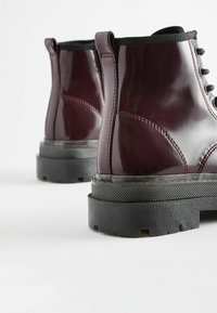 Bottines vernies bordeaux avec lacets noirs, semelle en caoutchouc et accents texturés. Elles présentent une finition lisse et une construction robuste.