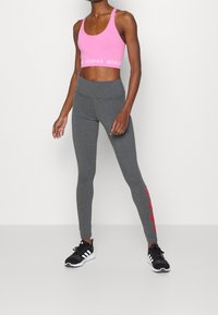 Rosa sport-bh med vit 'ADIDAS'-rem, parat med grå leggings som har röd märkning på vänster ben, bärs tillsammans med svarta träningsskor.