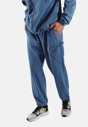 Persoon draagt blauwe Reebok joggingbroek met elastische boorden en bijpassend zip-up jasje, handen in de zakken, gecombineerd met witte en zwarte sneakers.