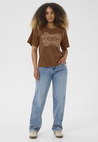 T-shirt marron à manches courtes avec un motif floral beige sur le devant, associé à un jean large bleu clair et des sneakers beiges.