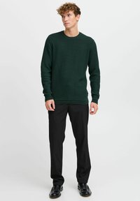 Pull en maille texturée vert foncé avec un col rond, des manches longues et des poignets côtelés, associé à un pantalon noir et des chaussures brillantes.