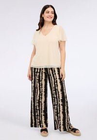 Donna con lungo capelli scuri, che indossa una maglietta beige a maniche corte e pantaloni ampi a righe neri e beige, sorridente e in piedi su uno sfondo bianco.