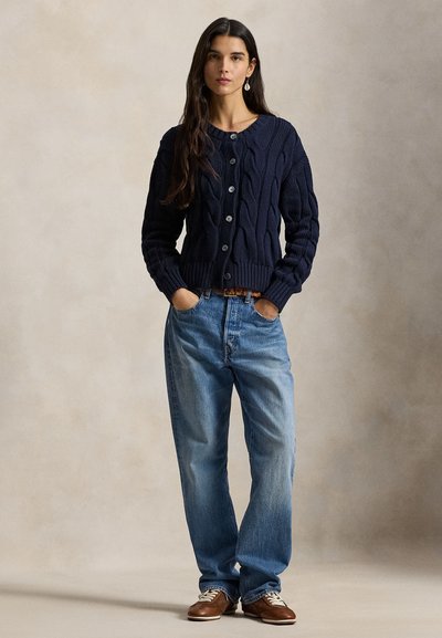 Πλεκτή ζακέτα ναυτικού στυλ με σχέδιο πλεξίδας, στρογγυλή λαιμόκοψη και κουμπιά, συνδυασμένη με ανοιχτόχρωμο τζιν και καφέ παπούτσια με κορδόνια.