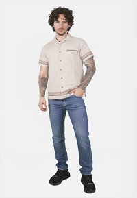 Camicia beige a maniche corte con dettagli ricamati, apertura con bottoni e taschino. Indossata con jeans blu e sneakers nere. Modello in posizione eretta.