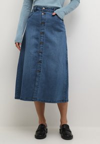 Gonna midi in denim blu con bottoni frontali, tasche a toppa e silhouette dritta. Indossata con scarpe slip-on nere.