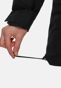 Giacca nera con polsini, caratterizzata da una mano che regola un cordino all'orlo, realizzata in un tessuto liscio e impermeabile.