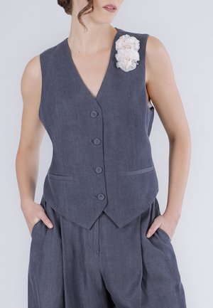 Mujer con chaleco sin mangas gris oscuro abotonado y broche floral blanco, pantalones holgados a juego, manos en los bolsillos, sobre fondo liso.