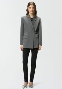 Blazer gris con diseño entallado, detalle de hebilla única, dos bolsillos frontales, combinado con leggings negros y zapatos planos metálicos.