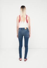 Vaqueros ajustados de color azul con cintura de altura media, bolsillos traseros y una etiqueta de cuero en la cinturilla. Camiseta blanca sin mangas con tirantes rojos. Sandalias planas.