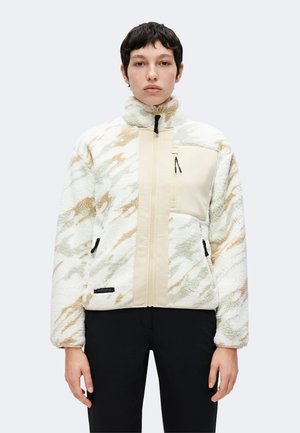 Personne portant une veste polaire beige et crème à motifs, avec une fermeture éclair frontale et une poche poitrine zippée, associée à un pantalon noir.