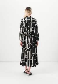 Femme aux longs cheveux blonds portant une robe noire avec des lignes abstraites blanches, nouée au dos, et des chaussures à petits talons noires, se tenant devant un fond blanc.