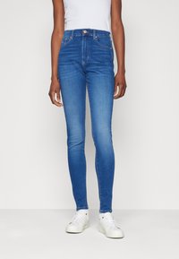 Vaqueros de denim azul de cintura alta con un corte ajustado, que cuentan con cinco bolsillos y un cierre de botón. Combinados con zapatillas blancas y una camiseta ajustada.