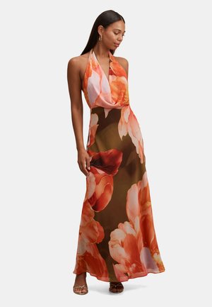 Nő ujjatlan, pánt nélküli hosszú ruhában, nagy narancssárga és rózsaszín virágmintákkal, nyitott orrú szandálokkal párosítva.