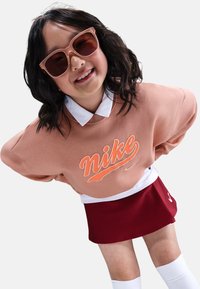 Sudadera color durazno de Nike con cuello blanco, con un logo naranja. Combinada con una falda roja plisada y calcetas blancas hasta la rodilla. Cabello castaño claro.