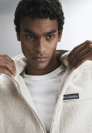 Junger Mann mit lockigem Haar, der eine cremefarbene Columbia-Fleecejacke über einem weißen Hemd vor einem einfarbigen Hintergrund anpasst.