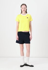 Geel katoenen T-shirt met een witte kraag en logo, gecombineerd met zwarte shorts. Model draagt witte sokken en zwarte instap schoenen.
