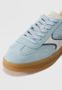 Jasnoniebieskie zamszowe sneakersy z okrągłym noskiem, kontrastującymi kremowymi akcentami, niebieskimi detalami i gumową podeszwą. Oferują płaskie sznurowadła i przeszywany wzór.