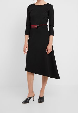 Frau trägt ein schwarzes asymmetrisches Midikleid mit Dreiviertelärmeln, einem roten Gürtel und schwarzen, hochhackigen Mules vor weißem Hintergrund.