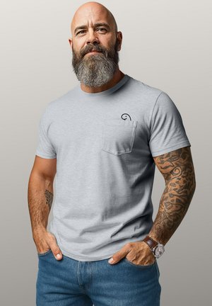 Bearded man with tattooed right arm wearing a light gray T-shirt and blue jeans, standing with hands in pockets against neutral background.