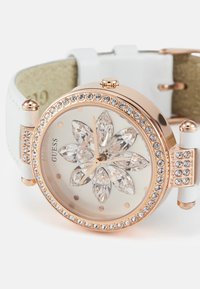 Roosa kuldne kell lillemustriga, millel on selged kristallaktsendid, valgest nahast rihm ja ringikujuline korpus, millel on sädelevad rhinestone'id.