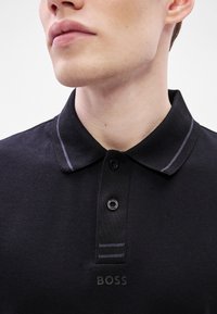 Melnas polo krekls ar teksturētu audumu, ar apkakli, trim pogām un kontrastējošu šuvumu, priekšpusē ar uzrakstu "BOSS".