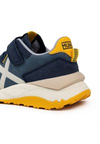 Sneaker blu navy con accenti gialli, materiali in camoscio e mesh, design a croce bianca prominente, suola bianca imbottita con gomma arancione testurizzata.
