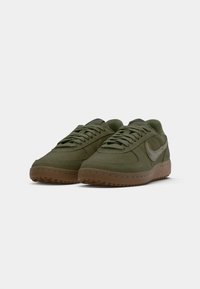 Par visoko maslinasto zelenih sukanih tenisica s teksturiranim bež Nike logom i smešim gumiranim potplatima na beloj pozadini.