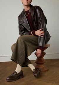Bruine leren loafers met een decoratieve band, textuurcrème sokken, olijfgroene wijde jeans en een oversized donkerbruine leren jas.