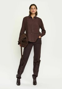 Chemise marron boutonnée avec une texture subtile, associée à un pantalon marron assorti et des bottes marron jusqu'aux genoux. Les accessoires comprennent un sac à épaule foncé.