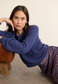 Marineblauer, gerippter Strickpullover mit strukturierten Ärmeln, kombiniert mit einem gemusterten Rock. Das Model lehnt an einem braunen Ledersessel. Neutrale Hintergrund.