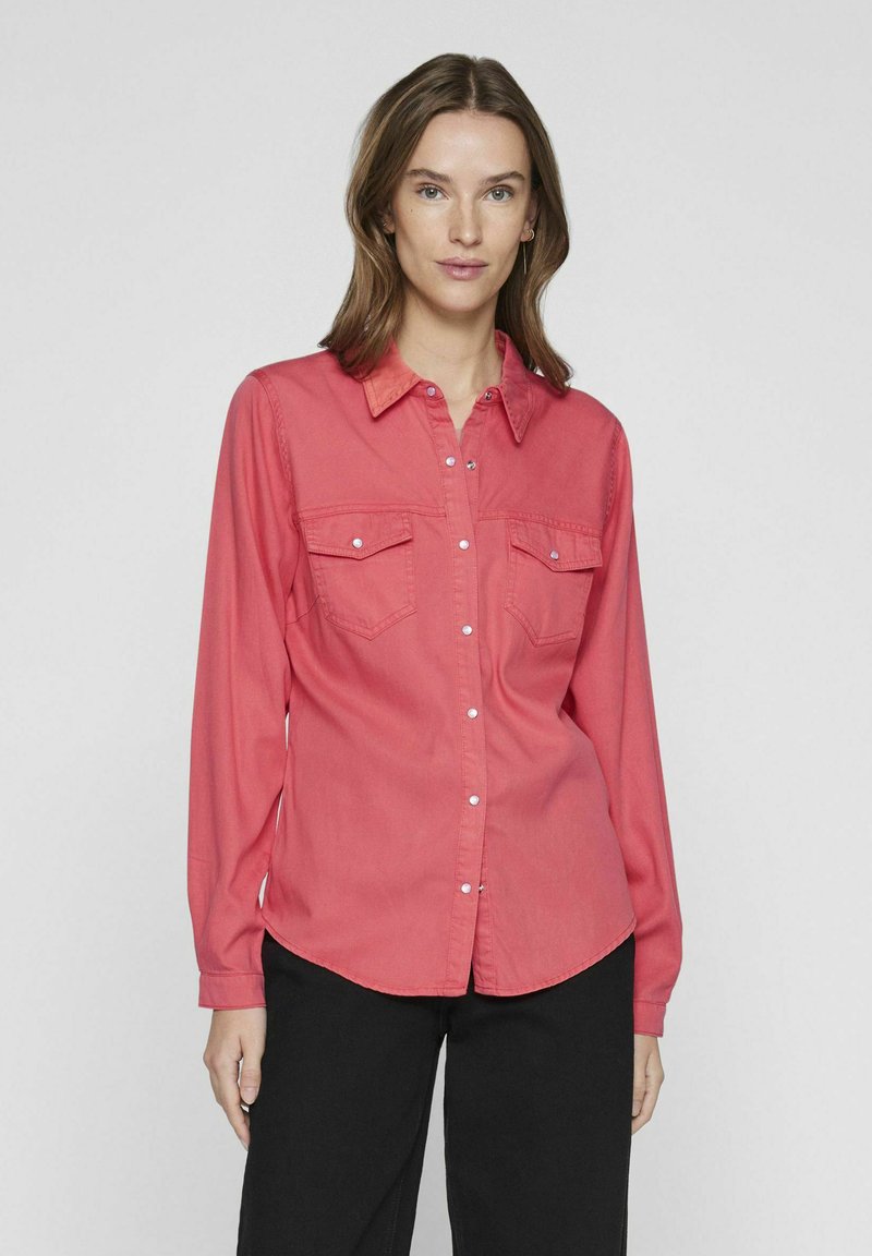 Camicetta a maniche lunghe color corallo con due tasche sul petto, design con colletto e una texture liscia, abbinata a pantaloni neri.