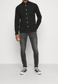 Camisa negra de botones con dos bolsillos en el pecho, mangas largas y cuello de camisa, combinada con jeans gris ajustados y zapatillas en blanco y negro.