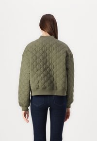 Veste bomber matelassée de couleur olive avec des poignets et un ourlet côtelés. Texture à motif en losanges notable, présentant une coupe décontractée et des épaules tombantes.