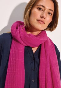 Fuchsia tørklæde med et struktureret ribbet design, lagt om halsen. Båret over en marineblå skjorte med knapper.