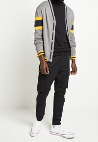 Cardigan gris avec des rayures jaunes et bleu marine, design boutonné, porté sur un col roulé noir. Associé à un pantalon cargo noir et des baskets blanches.