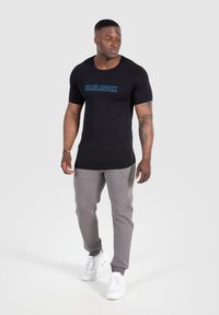 Camiseta negra ajustada con el logo azul "SMILODOX", combinada con joggers taper en gris claro y zapatillas deportivas blancas. Diseño simple y ajuste cómodo.