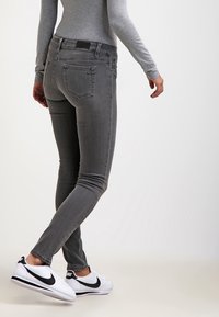 Osoba nosící šedé úzké džíny, šedý dlouhý rukáv a bílé tenisky Nike, stojící s překříženýma nohama před jednobarevným pozadím.