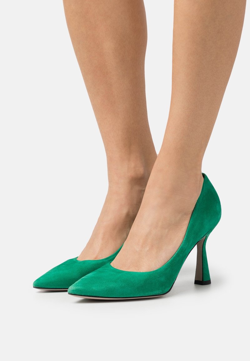 Escarpins en daim vert à bout pointu portés pieds nus, avec des talons fins mi-hauts effilés sur un fond neutre.