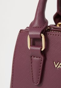 Piccola borsa color bordeaux realizzata in materiale sintetico testurizzato, con accenti in metallo dorato e un design strutturato. Presenta una chiusura a zip e manici robusti.