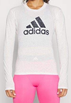 Zbliżenie na osobę noszącą białą, długorękawową koszulkę Adidas z czarnym logo oraz jasnoróżowe legginsy.