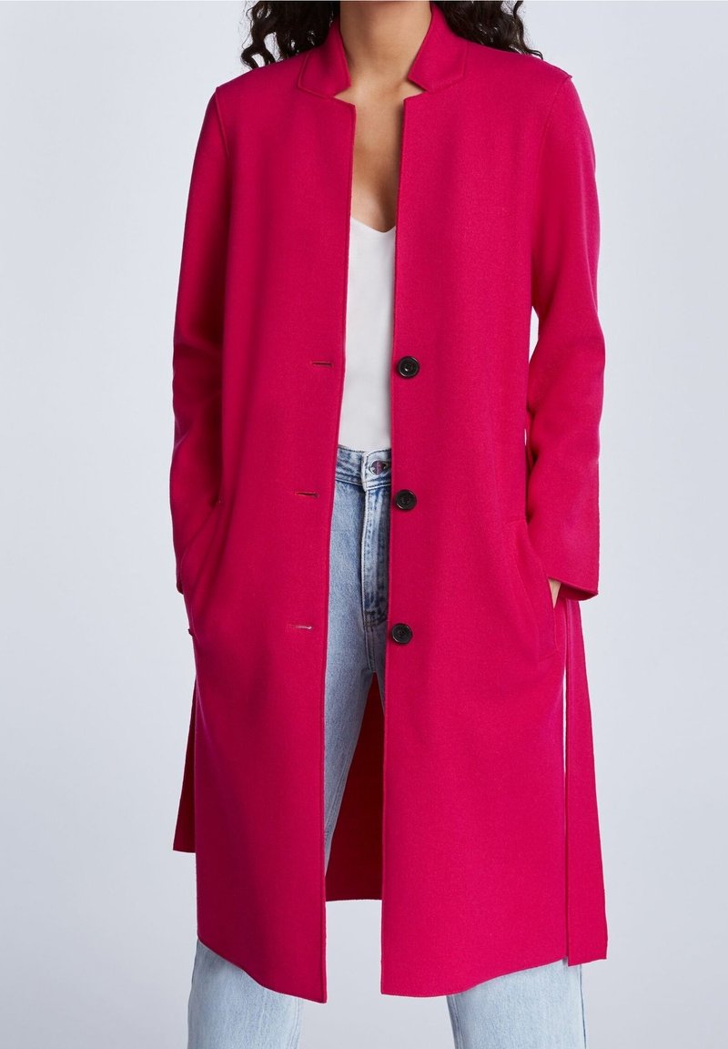 Fuksia pitkä takki, jossa on kaulus, avoin etuosa ja viisi mustaa nappia. Etutaskut ja sivuhalkiot lisäävät liikkumavapautta. Pehmeä, teksturoitu kangas.