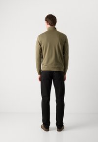 Maglione verde oliva con colletto alto, maniche lunghe e polsini a costine, abbinato a pantaloni neri e scarpe marroni, visto dalla parte posteriore.