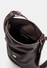 Braune Leder-Eimer-Tasche mit weicher Textur, silbernem Zubehör und einem kontrastierenden Nähgurt. Das Innere verfügt über eine Tasche und einen Stofffutter.