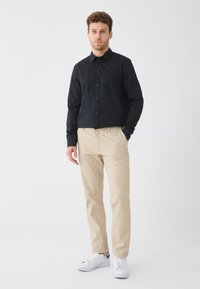 Mann steht vor weißem Hintergrund, trägt ein schwarzes Hemd mit Knöpfen, beige Hosen und weiße Sneakers, mit einer Hand in der Tasche.