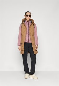 Gilet imbottito beige con cappuccio, indossato sopra un top rosa chiaro con zip, abbinato a pantaloni neri e sneakers beige. Il modello porta anche occhiali da sole.