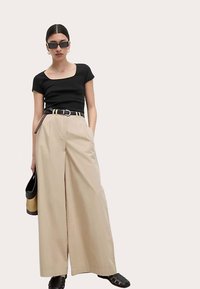 Sort top med firkantet halsudskæring, beige viddebenbukser, sort bælte og sorte slidesandaler; holder en rund, flettet taske. Minimalistisk design.