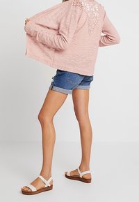 Světle růžový pletený cardigan s otevřeným designem, detail zad s květinovým výřezem, v kombinaci s denimovými šortkami a bílými sandály na dřevěné podrážce.