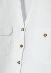 Chemise en lin blanc avec boutons en bois et deux poches poitrine à rabats, vue rapprochée montrant la texture du tissu et les détails de couture.