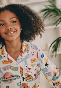 Chemise de pyjama colorée avec divers motifs de style cartoon, comprenant des billets de cinéma, des bonbons et des formes ludiques. Le matériau semble doux et léger.
